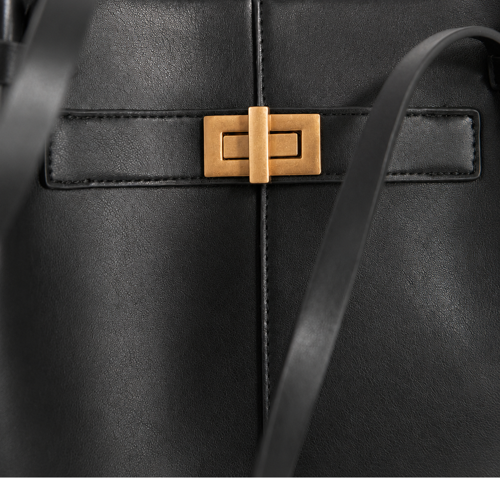 Close-up of a black leather handbag featuring a gold-tone rectangular turn-lock clasp and a matching black shoulder strap against a light off-white background.