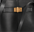 Close-up of a black leather handbag featuring a gold-tone rectangular turn-lock clasp and a matching black shoulder strap against a light off-white background.
